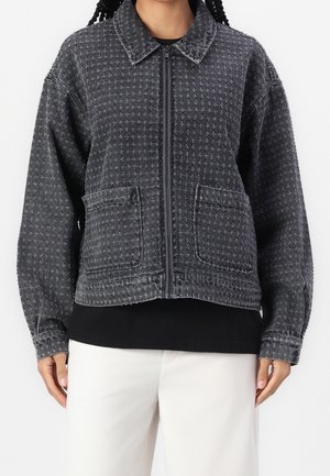 Femme portant une veste zippée gris foncé avec un motif croisé et des poches avant, associée à un pantalon blanc et un haut noir en dessous.