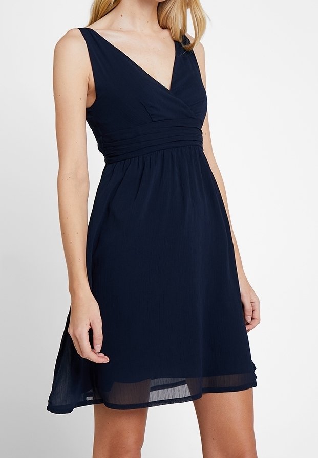 Vestido sin mangas azul marino con escote en V, cintura fruncida y dobladillo transparente a la altura de la rodilla, usado por una persona de pie.