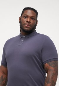 Kortermet polo-shirt i mørk lilla. Blødt, struktureret stof med tre-knaps lukning og ribbet krave. Fremhævet af synlige tatoveringer.