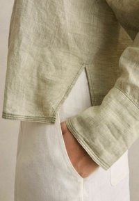 Olivgrünes Leinenoberteil mit einem cropped Design, das einen schrägen Saum und rohe Nahtdetails aufweist. Kombiniert mit weißen Leinenhosen und einer Seitentasche.