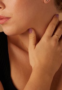 Nahaufnahme einer Hand mit heller Hautfarbe und einem goldenen Ring, die den Kiefer berührt. Die Nägel sind lavendel lackiert. Glatte Hautstruktur, dunkle Kleidung.