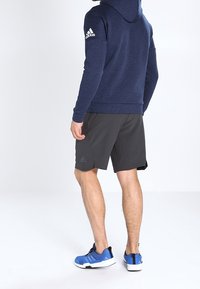 Felpa con cappuccio blu navy con finitura testurizzata, abbinata a pantaloni corti neri e scarpe da ginnastica blu. Logo Adidas sulla manica della felpa.