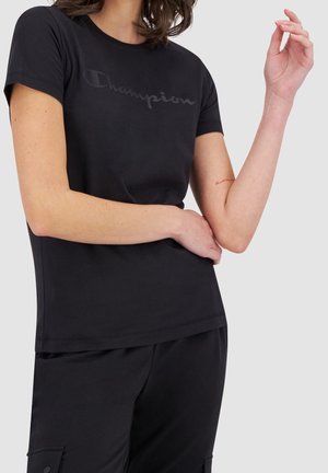 Kobieta w czarnej koszulce Champion i czarnych spodniach, z rękami skrzyżowanymi i uniesionymi na jednolitym jasnoszarym tle.