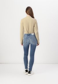 Beige ribbed sweater, slim-fit light blue jeans, white shoes with black accents, seen from the back against a plain background.