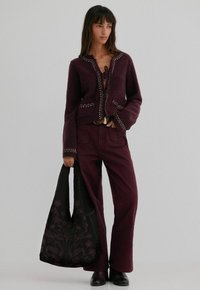 Bordeaux wollen cardigan met geborduurde randen, gecombineerd met bijpassende flare-broeken. Model heeft een zwarte tas met bloemenpatroon vast.