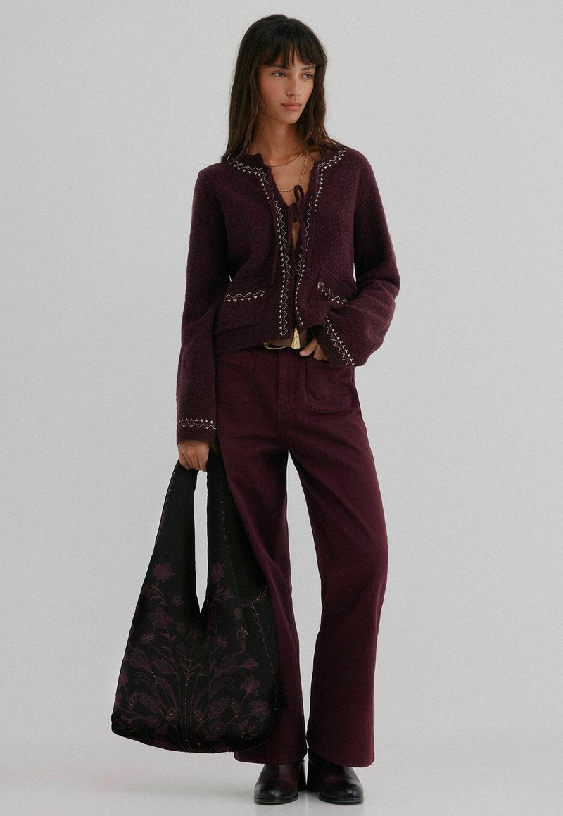 Bordeaux wollen cardigan met geborduurde randen, gecombineerd met bijpassende flare-broeken. Model heeft een zwarte tas met bloemenpatroon vast.