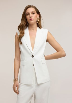 Frau mit langen Haaren, die einen ärmellosen weißen Blazer und passende weiße Hose trägt, ergänzt durch goldene Ohrringe und ein Armband, stehend vor neutralem Hintergrund.
