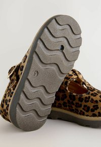 Chaussures imprimé léopard avec une palette de couleurs marron et noir, dotées d'une sangle à boucle et d'une semelle en caoutchouc grise texturée avec un motif ondulé.
