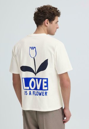 Muškarac okrenut u profil, nosi bijelu majicu s plavim cvijetnim motivom i tekstom "LJUBAV JE CVIJET" na leđima.