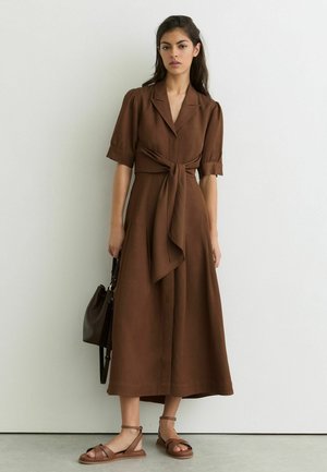 Kobieta stojąca przy białej ścianie, ubrana w brązową sukienkę midi z wiązaniem w talii, trzymająca ciemnobrązową torebkę i mająca na stopach brązowe sandały.