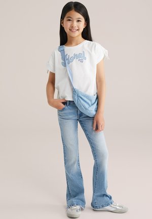 Weißes Baumwoll-T-Shirt mit blauer Blumenstickerei; hellblaue, ausgestellte Jeans; kleine hellblaue Umhängetasche; graue Sneakers.