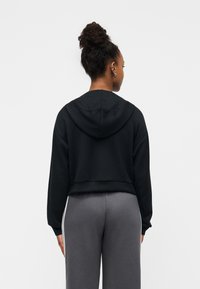 Κοντό μαύρο φούτερ με κουκούλα και μακριά μανίκια, με απαλή υφή. Συνδυάζεται με γκρι παντελόνι που έχει πλευρικές ρίγες.