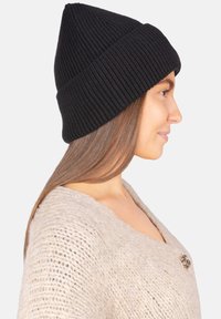 Bonnet en maille côtelée noire avec une pointe effilée ; texture douce ; porté avec un pull en maille beige clair orné d'un bouton décoratif.
