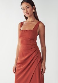Ein rostfarbendes Kleid mit quadratischem Ausschnitt, breiten Trägern und gerafften Details an der Seite, aus einem glatten Material gefertigt.