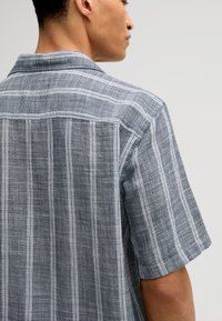 Grijs gestreepte kortemouwen shirt met een kraag. Gemaakt van een textuurstof met een patroon van dunne witte strepen tegen een donkerder achtergrond.