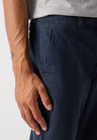 Nahaufnahme einer Männerhand, die neben einer Hose in Marineblau ruht, trägt ein hellgraues Hemd, mit sichtbaren Armvenen und einem teilweisen Tattoo.