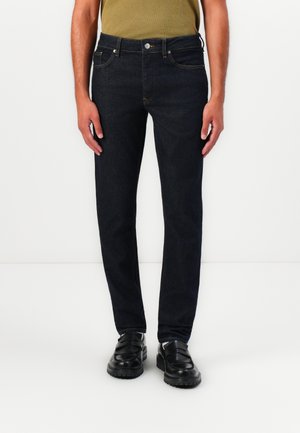 Jean en denim bleu foncé avec une coupe slim, présentant un design classique à cinq poches et des coutures couleur cuivre. La ceinture inclut une fermeture à bouton.