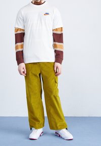 Nike vit t-shirt med blå logotyp, kombinerad med bruna långärmade tröjor med ränder. Oljefärgad kordbyxbyxor och vita sneakers.