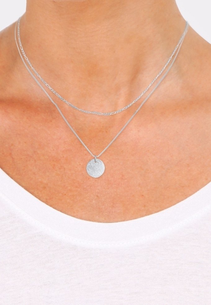 Deux colliers en argent autour du cou d'une personne, l'un avec une chaîne fine et l'autre avec un pendentif circulaire, sur une chemise blanche.