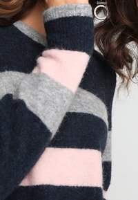 Osoba v načechraném svetru s horizontálními námořnickými, šedými a růžovými pruhy a s částečně viditelnými kudrnatými hnědými vlasy.