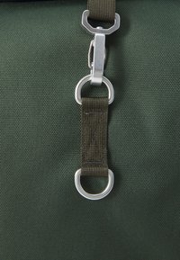 Sac en tissu vert avec un clip en métal argenté et une sangle en tissu vert foncé avec des détails de couture, attachée à un anneau circulaire.