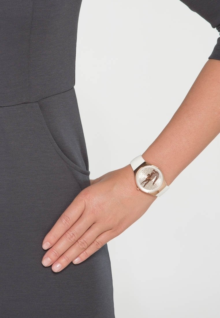 Main féminine reposant sur la hanche portant une montre-bracelet blanche avec un boîtier doré et un cadran scintillant.