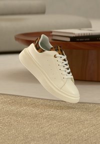 Λευκό αθλητικό παπούτσι με λεπτομέρειες λεοπάρ στο γλώσσα και στον γιακά, αιωρούμενο πάνω από μπεζ χαλί σε ένα μοντέρνο σαλόνι.