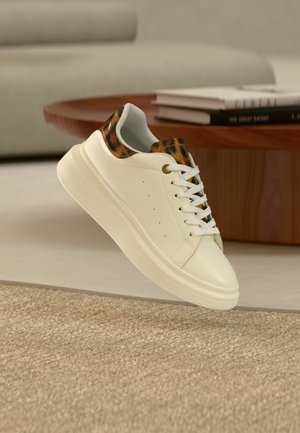 Baskets blanches avec des accents imprimé léopard sur la languette et le col, flottant au-dessus d'un tapis beige dans un salon moderne.