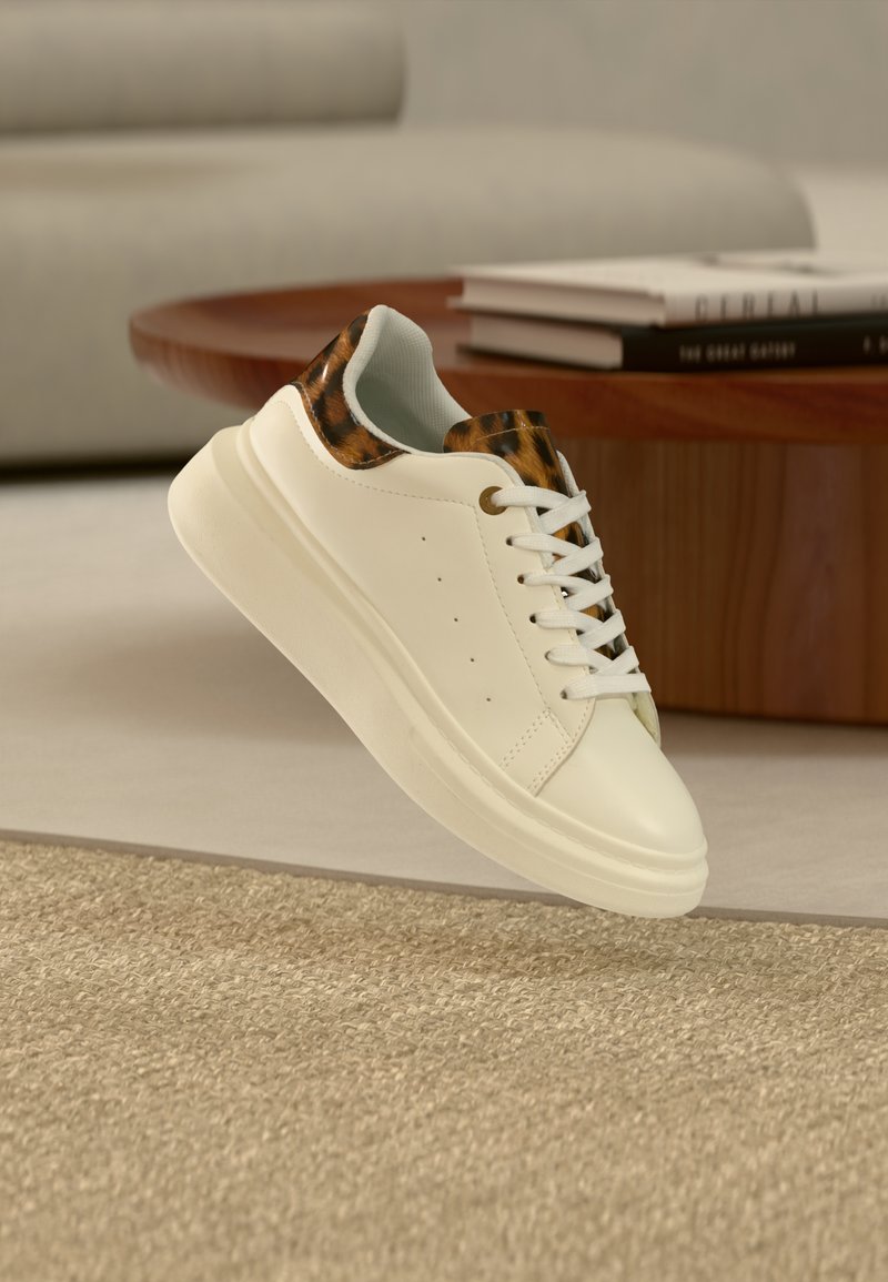 Λευκό αθλητικό παπούτσι με λεπτομέρειες λεοπάρ στο γλώσσα και στον γιακά, αιωρούμενο πάνω από μπεζ χαλί σε ένα μοντέρνο σαλόνι.
