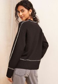 Maglione nero in filato testurizzato, con cuciture bianche lungo le spalle e orlo a coste. Indossato con jeans grigi chiari.