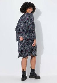 Ein schwarzes Kleid mit einem bunten abstrakten Muster, ausgestellten Ärmeln und einem Rüschensaum. Das Model kombiniert es mit schwarzen Schnür-Ankle Boots.