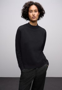 Černý svetr s vysokým výstřihem a dlouhými rukávy. Měkká textura, tvarující silueta s mírným nabíráním na ramenou. V kombinaci s tmavými vzorovanými kalhotami.