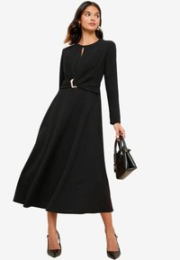 Schwarzes langärmliges Kleid mit einem Schlüsselloch-Ausschnitt und gedrehtem Bund, der mit einem metallischen Akzent versehen ist. Das Modell hält eine kleine schwarze Handtasche.