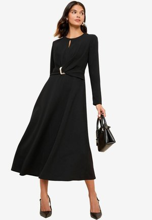 Black long-sleeve dress with a keyhole neckline and twisted waist featuring a metallic accent. Model holds a small black handbag.