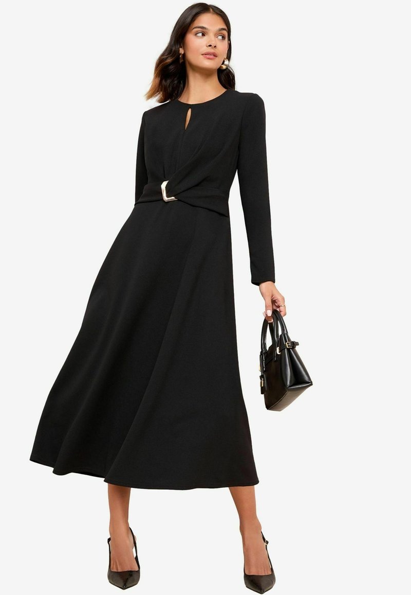 Schwarzes langärmliges Kleid mit einem Schlüsselloch-Ausschnitt und gedrehtem Bund, der mit einem metallischen Akzent versehen ist. Das Modell hält eine kleine schwarze Handtasche.