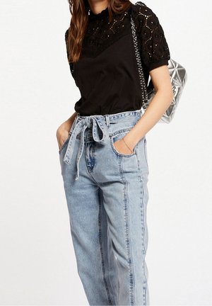Woman wearing black lace short-sleeve top, high-waisted light blue jeans with tied belt, and carrying a silver quilted shoulder bag.