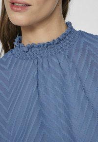 Niebieska bluzka z falbanistym dekoltem, teksturowanym wzorem w zygzaki oraz lekkim materiałem, prezentująca delikatny i zorganizowany design.