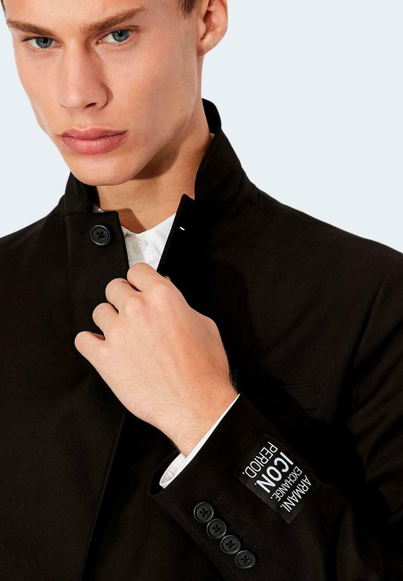 Joven ajustando el cuello de una chaqueta negra de Armani Exchange con una camisa blanca debajo, visible el logo de Armani Exchange en la manga.