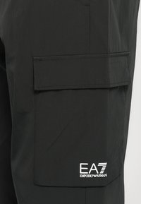 Melni laikapārnēsājamie bikses, kas izgatavotas no viegla auduma, ar lielu sānu kabatu un balto diegu "EA7" logo kreisajā pusē.