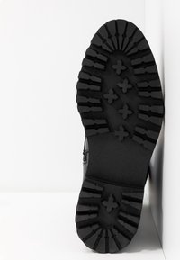 Semelle d'une botte noire avec des motifs de semelle profonds et robustes comprenant des barres horizontales et des formes en croix, appuyée contre un mur blanc.