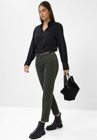 Schwarzes Langarmshirt, grüne Cordhosen, schwarze Stiefeletten mit dicken Sohlen und eine kleine schwarze Handtasche mit Trägern.