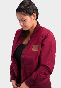 Burgundy bomber jacket with a ribbed collar and cuffs, featuring a leather patch on the chest and a zip closure. Smooth, lightweight fabric.