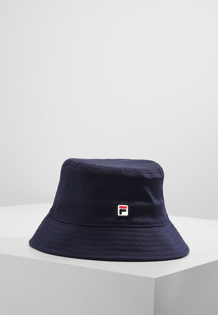 Chapeau seau bleu marine avec un petit écusson carré orné d'un logo rouge, blanc et noir sur le devant, posé sur une surface blanche contre un fond clair.