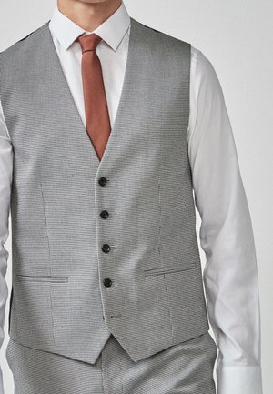 Homme portant une chemise blanche, une cravate couleur rouille et un gilet boutonné à carreaux gris avec un pantalon assorti.