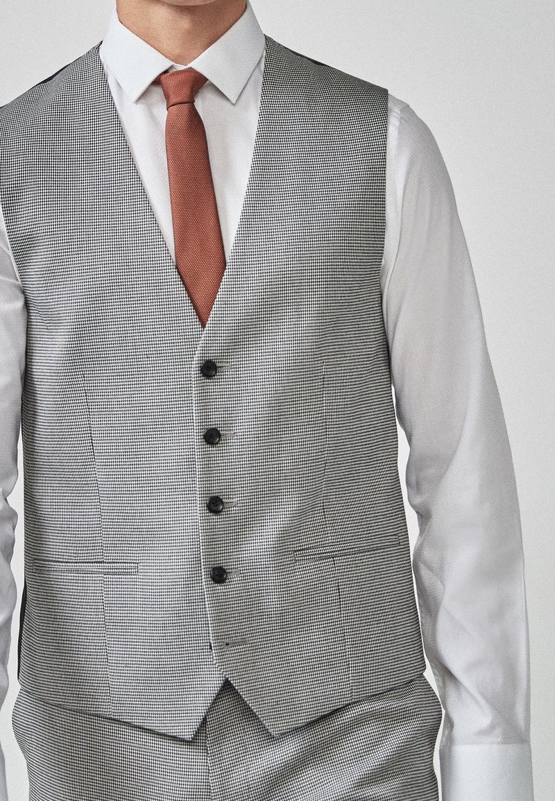 Homme portant une chemise blanche, une cravate couleur rouille et un gilet boutonné à carreaux gris avec un pantalon assorti.
