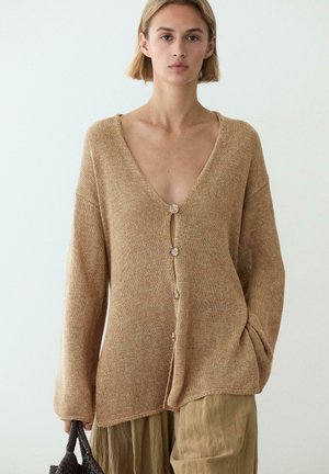 Donna con capelli biondi corti che indossa un cardigan lavorato a maglia largo color sabbia con bottoni e pantaloni larghi marrone chiaro, tenendo una borsa intrecciata scura.