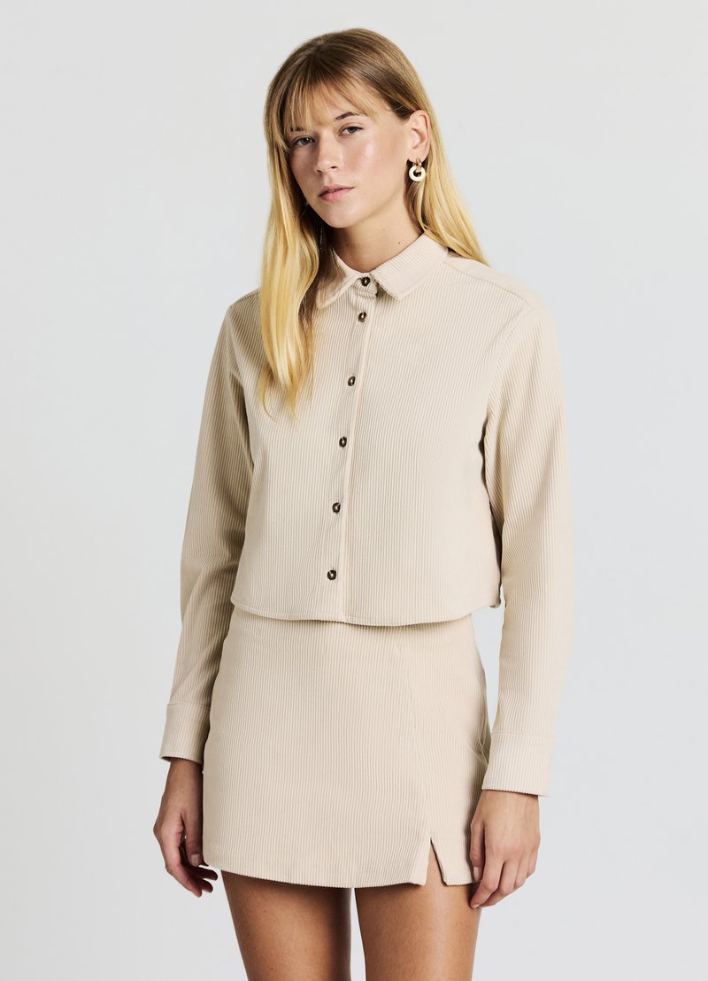 Blusa beige a coste con colletto e bottoni sul davanti, abbinata a una minigonna a coste coordinata con spacco laterale. Texture liscia.