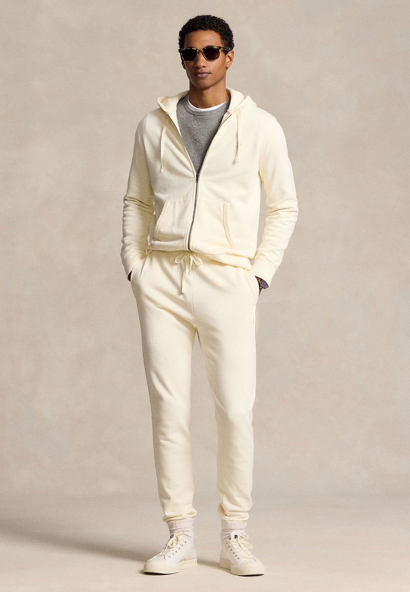 Camisola com fecho e capuz em tom creme, com cordão, combinada com calças de corrida a condizer. Vestida por cima de uma t-shirt cinzenta, complementada com ténis brancos e óculos de sol.