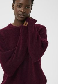 Pull en maille bordeaux avec un col rond, coupe ample et manches longues. Texture douce avec une légère fuzziness. Aucun motif ni élément métallique visible.