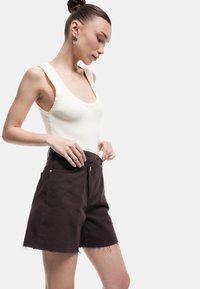 Pantalones cortos marrones con dobladillo sin terminar, bolsillos frontales y detalles dorados, combinados con una camiseta de tirantes de color crema con detalles en forma de concha.